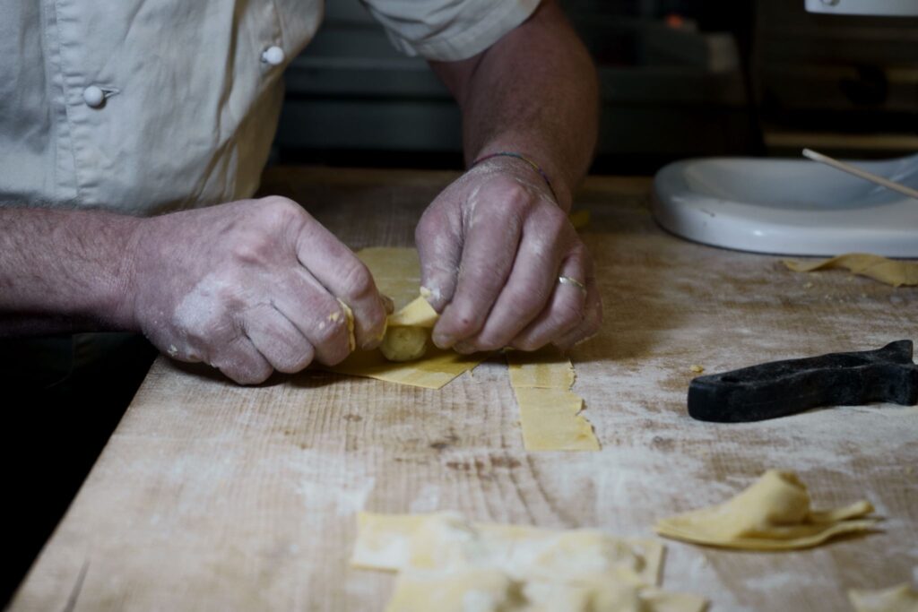 Hand-Shaping Fresh Pasta Dough Close-up of a chef's flour-dusted hands shaping yellow pasta dough on a wooden table, with a pasta cutter and rolled dough in the background.