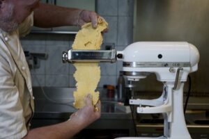 A chef rolling yellow pasta dough through a KitchenAid machine attachment, with flour-dusted hands and a kitchen counter in the background.