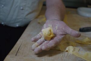 A chef's flour-covered hand holding a folded piece of yellow pasta dough, with more dough pieces on a wooden table.