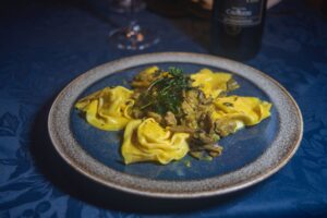 A blue plate with yellow pasta topped with a mushroom and herb sauce, set on a dark blue tablecloth with a wine bottle in the background.