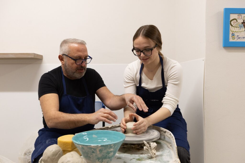 Two people shaping clay on a pottery wheel in a Tuscan ceramics masterclass, with tools and a finished bowl nearby.