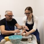 Two people shaping clay on a pottery wheel in a Tuscan ceramics masterclass, with tools and a finished bowl nearby.
