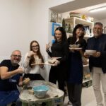 A group of students holding their pottery creations during a Tuscan ceramics masterclass, with a pottery wheel and shelves in the background.