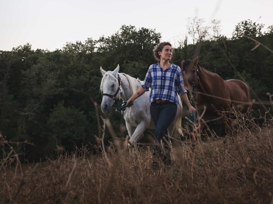Horse guide Margherita walking a white and a chestnut horse through a dry Tuscan field — horseback riding in Italy