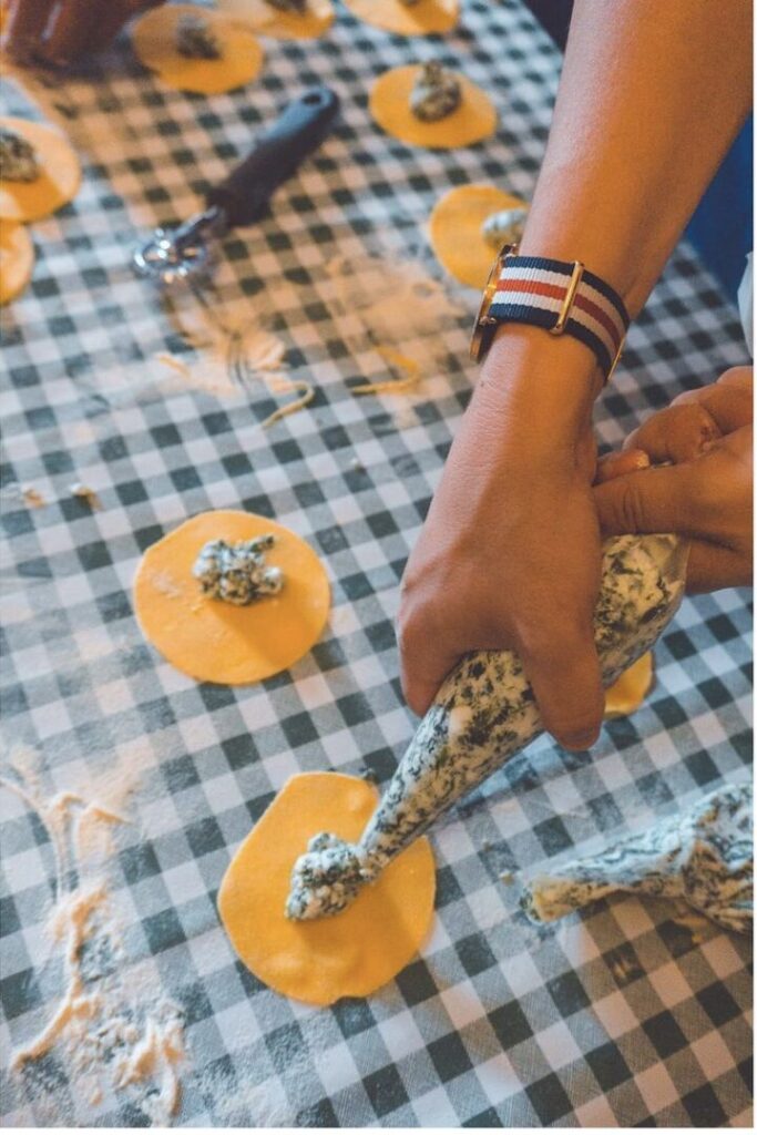 Hands piping green ravioli filling onto pasta circles on a checkered cloth, a cooking class with kids Tuscany can join.