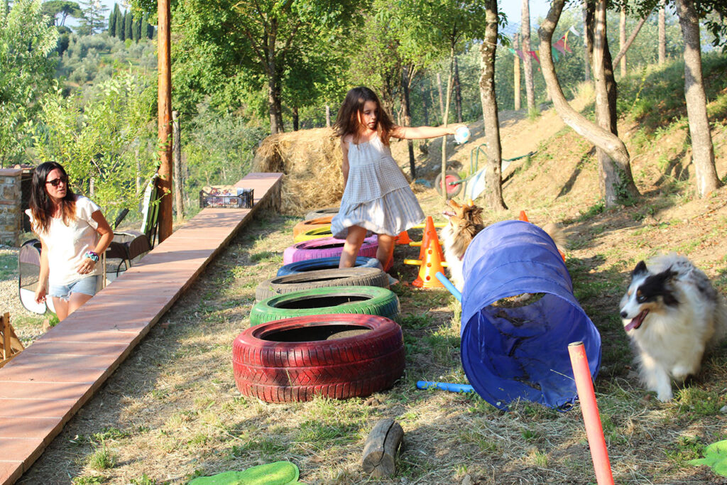 Girl crossing painted tires beside a play tunnel and two dogs, things to do in Tuscany with kids at a countryside farm.