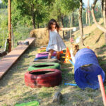 Girl crossing painted tires beside a play tunnel and two dogs, things to do in Tuscany with kids at a countryside farm.